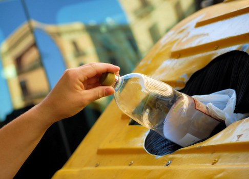A person's hand is shown holding a small glass bottle filled with clear liquid, with the bottle angled slightly downward. The background features a yellow vehicle, likely a van or truck, with a rounded body and a black circular opening on its side, possibly where rubbish is loaded or unloaded. The vehicle's surface appears shiny and smooth, with visible reflections of nearby buildings and sky, indicating bright outdoor lighting. The scene is set on a paved street or driveway, with a blurred urban environment in the distance, including structures with windows and façades that suggest a residential or commercial area in Bromley. The image context relates to waste disposal or rubbish removal, fitting with services provided by Office Clearance Bromley in the local area. The focus on the hand and bottle emphasizes a detail-oriented approach, relevant to environmentally conscious waste management practices within the community.