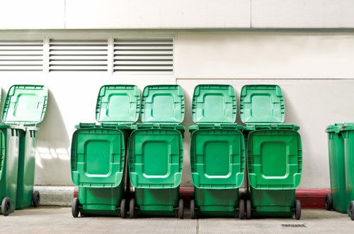 A row of six identical green wheeled rubbish bins with closed lids, positioned on a paved area in front of a white building with ventilation louvers near the roofline. The bins are made of durable plastic with textured surfaces and are arranged in two rows of three, evenly spaced alongside each other. The environment appears to be an outdoor service or storage area, potentially adjacent to a commercial or industrial building. The background features a smooth, light-colored wall with horizontal ventilation slats, and the ground surface is clean and level. The scene reflects a typical waste collection or rubbish disposal setting, consistent with services offered by companies such as Office Clearance Bromley, serving the Bromley area and surrounding postcode districts.