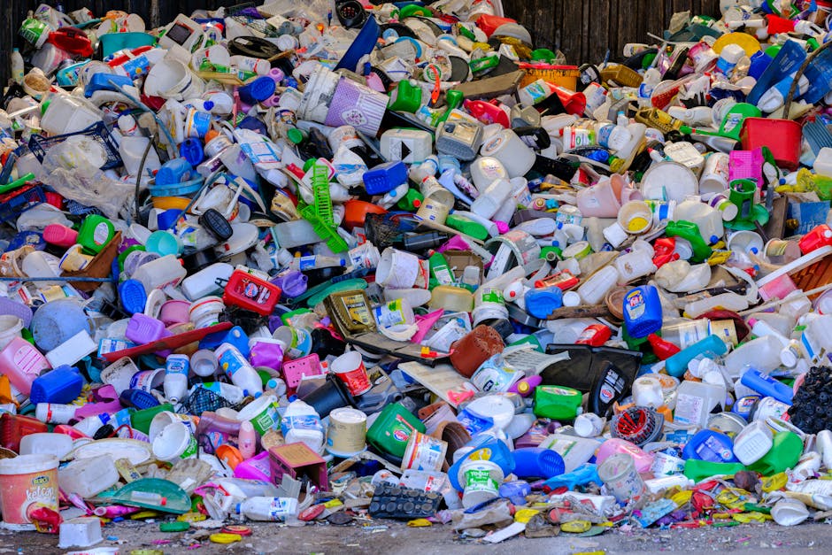 A row of six large wheelie bins made of plastic, positioned on a paved surface in front of a tall, rectangular shipping container. The bins are predominantly white with slightly weathered surfaces and