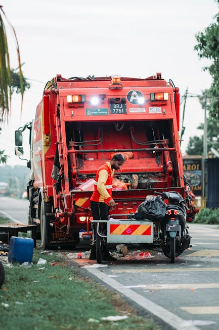 A sanitation worker dressed in a bright yellow and red uniform is seen operating a red rubbish collection vehicle on a street. The worker is leaning forward, engaged in clearing or maintaining the rea