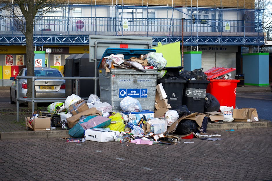 A commercial waste collection truck parked on a city street during daytime, with the truck’s large, rectangular metal container filled with collected rubbish visible in the foreground. The container